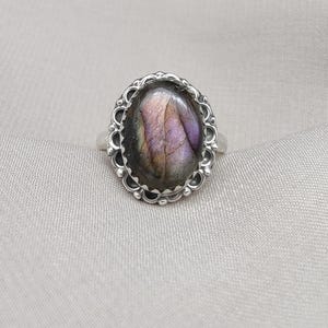 Puede incluir: Un anillo de plata con una piedra preciosa de labradorita ovalada. La piedra preciosa muestra tonos morados, azules y verdes. El anillo tiene un engaste decorativo con bisel festoneado. El anillo está sobre un fondo neutro.