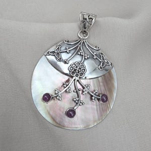 Puede incluir: Colgante de plata con una gran base redonda e iridiscente de nácar. La mitad superior es plateada con un diseño floral, y la mitad inferior tiene tres pequeñas piedras preciosas redondas y moradas. El colgante tiene una argolla decorativa.
