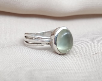 May include: A silver ring with a light green, oval-shaped gemstone. The ring has a split band design with three parallel lines on each side of the gemstone. The gemstone is smooth and polished, set in a silver bezel.