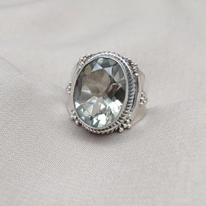 Puede incluir: Un anillo de plata con una gran piedra preciosa ovalada de color verde pálido. El anillo tiene una banda de plata ornamentada con detalles decorativos alrededor de la piedra y en los laterales. El anillo se muestra sobre una tela gris claro.