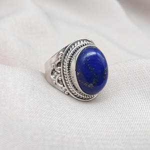 以下が含まれることがあります： 大きな楕円形の濃い青色の宝石をあしらったシルバーリング。 宝石は、石の周りとリングの側面に複雑なディテールが施された装飾的なシルバーバンドにセットされています。 リングは明るい色の生地の上に表示されています。