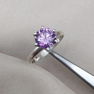 May include: A silver ring featuring a large, round, light purple gemstone. The gemstone is held in place by six prongs. The ring is displayed on a light gray surface.