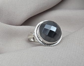 Puede incluir: Un anillo de plata con una gran piedra preciosa negra facetada. La piedra preciosa redonda está engastada en un bisel de plata decorativo con un diseño de cuerda trenzada. El anillo se muestra sobre una tela de tono neutro.