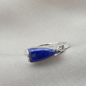 Pode incluir: Um anel de prata com uma grande pedra preciosa azul escura facetada. A pedra preciosa é retangular com uma extremidade ligeiramente arredondada. A banda do anel é fina e feita de prata, com um elemento decorativo perto da pedra.