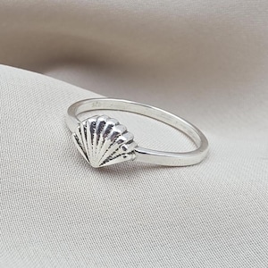 May include: A silver ring featuring a small, detailed seashell motif centred on a thin, polished band. The metal surface has a bright, reflective finish with visible ridges on the shell design and a small 925 hallmark stamped on the inner band.