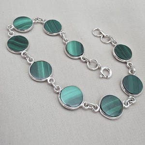 Pode incluir: Uma pulseira de prata com pedras circulares de malaquite. As pedras verdes são fixadas numa corrente de prata com um fecho. A pulseira é exibida sobre um fundo neutro.