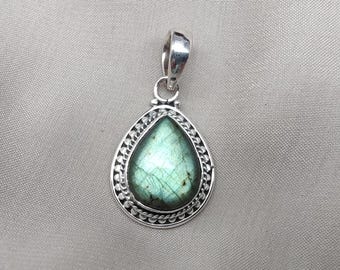 May include: A silver pendant with a teardrop-shaped labradorite stone. The stone has a green and blue iridescent sheen. The pendant is framed by a decorative silver border with a beaded design. The bail is a simple, polished silver loop.