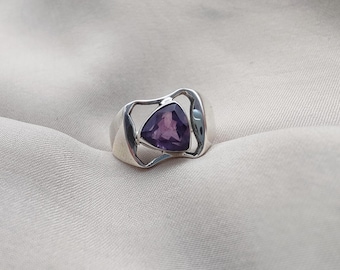 May include: A silver ring with a triangular, purple gemstone. The ring has an open, modern design with a polished finish. The gemstone is set in a bezel setting, and the ring is displayed on a neutral-colored fabric.