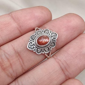 Op de afbeelding: Een zilveren ring met een ovale, rood-oranje edelsteen in het midden. De edelsteen is omgeven door een sierlijke, bloem geïnspireerde zetting. De ring heeft een eenvoudige, dunne band.