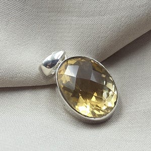 Puede incluir: Un colgante de forma ovalada con una piedra preciosa translúcida de color marrón dorado facetada, engastada en un marco de metal plateado pulido. El colgante incluye una anilla plateada lisa y redondeada en la parte superior.