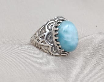 May include: A silver ring with a large, oval, pale blue gemstone. The ring's band has an intricate, engraved pattern. The gemstone is held in a bezel setting. The ring is shown on a neutral-toned background.
