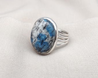 May include: Silver ring featuring an oval k2 stone. The stone has a mix of blue, white, and black patterns. The ring's band has a multi-strand design. The ring is set against a white fabric background.