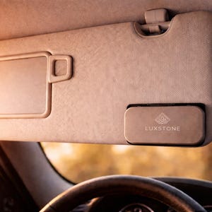 May include: A beige car sun visor with a rectangular storage compartment. The visor is attached to the car's ceiling and has a small, rectangular mirror. A rectangular accessory with the word "LUXSTONE" is attached to the visor.