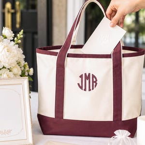 Könnte beinhalten: Cremefarbene Tragetasche mit bordeauxfarbenen Akzenten und dem Monogramm "JMB". Die Tasche hat bordeauxfarbene Griffe und einen Boden. Eine Person legt eine Karte in die Tasche. Ein gerahmtes Bild, Blumen, Kerzen und ein Umschlag befinden sich ebenfalls auf dem Tisch.
