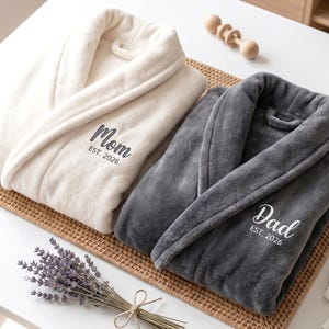 May include: Two plush bathrobes, one cream and one gray, are displayed on a woven mat. The cream robe has "Mom EST. 2026" embroidered on it, while the gray robe says "Dad EST. 2026". A bundle of lavender and baby shoes are also visible.
