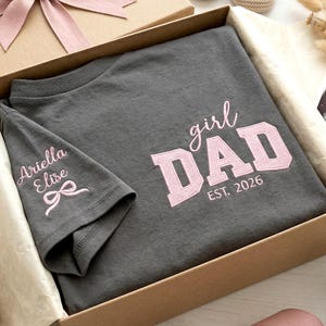 May include: A charcoal gray t-shirt folded inside a cardboard gift box. The shirt features light pink embroidered text that reads girl DAD EST. 2026 on the front and the name Ariella Elise with a small bow graphic on the sleeve.