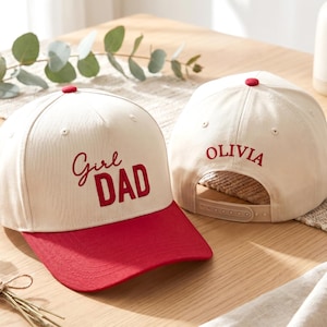 May include: Two off-white baseball caps with red brims and red buttons on top. One cap features the red embroidered text Girl DAD on the front. The other cap is shown from the back, displaying the red embroidered name OLIVIA above the strap.