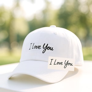 Op de afbeelding: Een witte baseballpet met de geborduurde tekst "I Love You" in zwart script. Een kleine kaart met dezelfde zin is voor de pet geplaatst. De achtergrond is een vage buitenscène.