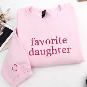 A light pink sweatshirt featuring “favorite daughter” embroidery across the chest.
The lettering is stitched in deep rose thread, with a small matching heart detail on the sleeve.
Sweet, playful, and perfect for everyday wear.