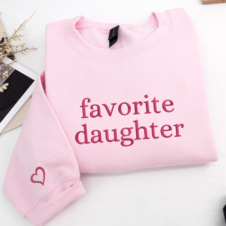 A light pink sweatshirt featuring “favorite daughter” embroidery across the chest.
The lettering is stitched in deep rose thread, with a small matching heart detail on the sleeve.
Sweet, playful, and perfect for everyday wear.