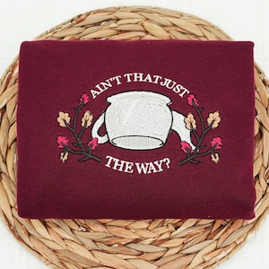 May include: A burgundy sweatshirt with the embroidered phrase "Ain't That Just The Way?" above a white teapot design. The design includes floral accents in pink and brown, set against a woven background.