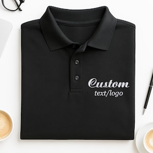 Könnte beinhalten: Schwarzes Poloshirt mit Kragen und drei Knöpfen. Der Schriftzug "Custom text/logo" ist in Weiß aufgestickt. Das Shirt ist sauber auf einer weißen Oberfläche gefaltet.
