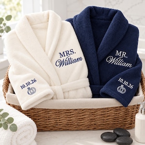 May include: A cream-colored bathrobe embroidered with 'MRS. William' and a navy blue bathrobe embroidered with 'MR. William'. Both robes feature the date '10.10.26' and an icon of two interlocking rings on the sleeve cuffs.