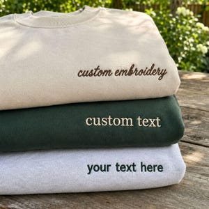 May include: A stack of three crewneck sweatshirts in beige, dark green, and light gray. The beige sweatshirt features brown embroidered text reading custom embroidery. The dark green sweatshirt has beige text reading custom text. The gray has dark text.