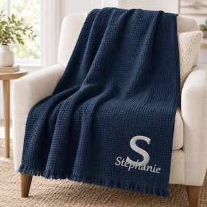 May include: A navy blue waffle-weave throw blanket featuring a large embroidered letter S and the name Stephanie in white thread. The blanket has a textured grid pattern and a fringed edge, draped over a light-colored upholstered armchair.