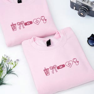 Könnte beinhalten: Zwei hellrosa Sweatshirts mit bordeauxfarbenen Motiven: ein Getränk, eine Schleife, ein Pflaster, ein Herz mit Kreuz und ein Stethoskop. Auf einer weißen Oberfläche, mit Kamera und Sonnenbrille.