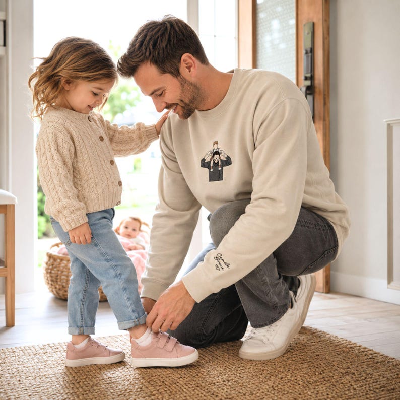 May include: A beige sweatshirt with a graphic of a person carrying a child on their shoulders. The sweatshirt is worn by a person kneeling to help a child with pink shoes. The child wears a beige cardigan and blue jeans.