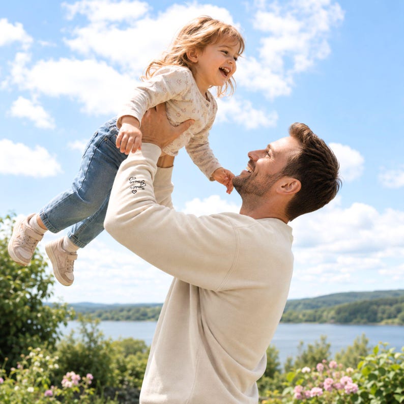 May include: A light beige sweatshirt with embroidered text on the sleeve. A child is being held up in the air, wearing a floral print top, blue jeans, and pink shoes. The background features a blue sky and green foliage.