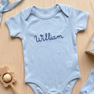 Op de afbeelding: Een lichtblauwe babybody met de naam "William" geborduurd in marineblauwe letters. De body met korte mouwen heeft drukknoopsluitingen. Een houten rammelaar en een geschenkdoos met blauw lint zijn ook zichtbaar.
