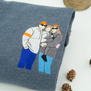 A Dark Heather sweatshirt featuring a custom embroidered family portrait on the left chest.
The design uses multi-color thread for clothing and hair details, creating a bold and modern look.