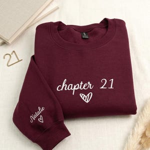 以下が含まれることがあります： 白の刺繍で「chapter 21」とハートのデザインが施された、マルーン色のスウェットシャツ。袖には「Natalie」という名前とハートが刺繍されています。スウェットシャツは白い表面に折りたたまれています。