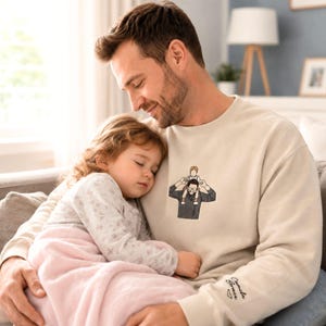 May include: A beige sweatshirt with a graphic of a man carrying a child on his shoulders. The sweatshirt is worn by a man holding a sleeping child. The sleeve has embroidered text. The scene is set indoors.