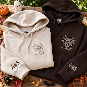 May include: A sand-coloured hoodie and a dark brown hoodie, both featuring a heart-shaped spider web embroidery on the chest with the Roman numeral date VII.XVII.XXIV below it. The sleeves are embroidered with the letters A and S with a spider.