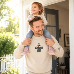 May include: A beige sweatshirt with a graphic of a person carrying a child on their shoulders. The sweatshirt is worn by a person with a child on their shoulders. The child is wearing a light-colored top and jeans.