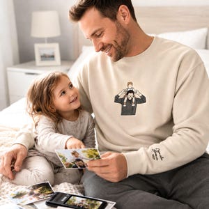 May include: A cream-colored sweatshirt with an embroidered illustration of a person with a child on their shoulders. The sweatshirt has the name "Camila Grace" embroidered on the sleeve. A person and a child are looking at photos.