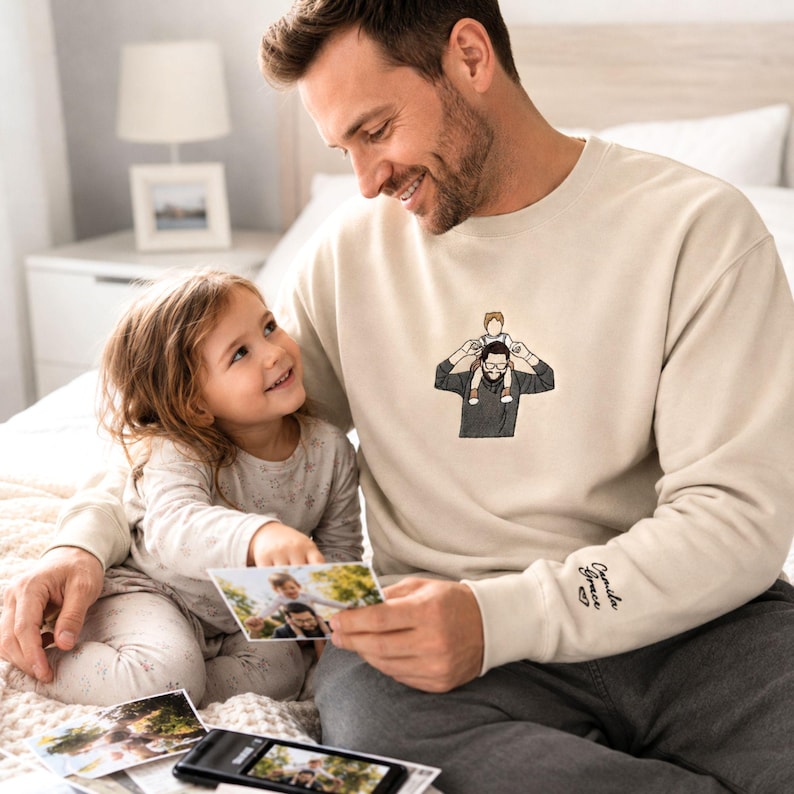 May include: A cream-colored sweatshirt with an embroidered illustration of a person with a child on their shoulders. The sweatshirt has the name "Camila Grace" embroidered on the sleeve. A person and a child are looking at photos.