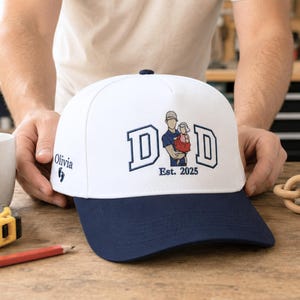 May include: A white baseball cap with a navy blue brim featuring embroidered text that reads DAD and Est. 2025. The design includes an illustration of a person holding a child. The side of the cap features the name Olivia and two small footprints.