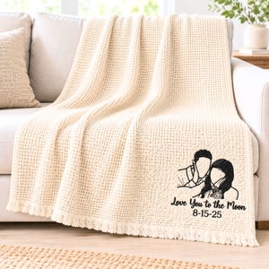 May include: A cream-colored waffle-knit throw blanket with fringe edges. The blanket features black embroidery of a couple's outline, a small floral detail, and the text "Love You to the Moon" followed by the date "8-15-25" in a script font.