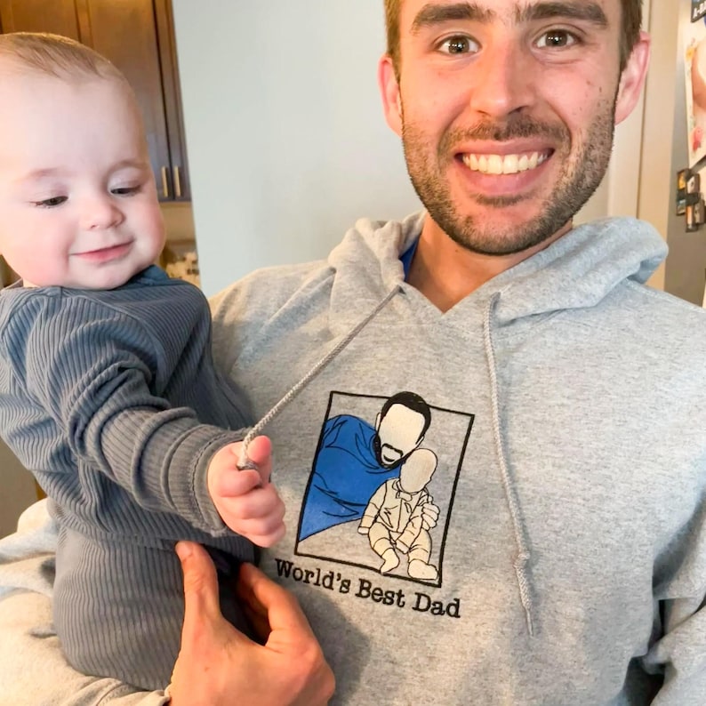 A Sport Grey hoodie featuring a custom embroidered dad-and-baby portrait on the middle chest, stitched in multi-color thread.
Below the artwork, the phrase “World’s Best Dad” is embroidered in black thread.