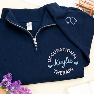 Puede incluir: Sudadera azul marino con cremallera de cuarto de cremallera y cremallera blanca. La sudadera presenta el texto "OCCUPATIONAL Kaylie THERAPY" en un diseño circular, un estetoscopio y dos corazones. La manga tiene un estetoscopio bordado.
