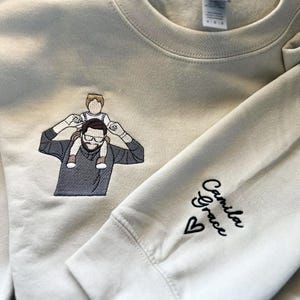 A sand sweatshirt featuring a custom embroidered dad-and-child portrait on the right chest using multi-color thread.
The left sleeve is personalized with the name “Camila Grace” and a small heart in black thread.