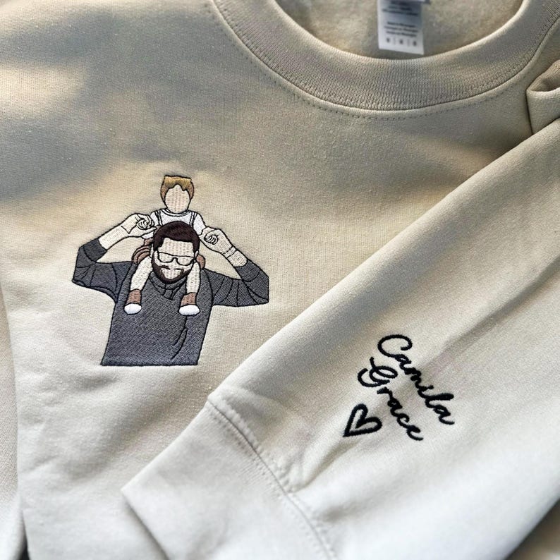 A sand sweatshirt featuring a custom embroidered dad-and-child portrait on the right chest using multi-color thread.
The left sleeve is personalized with the name “Camila Grace” and a small heart in black thread.