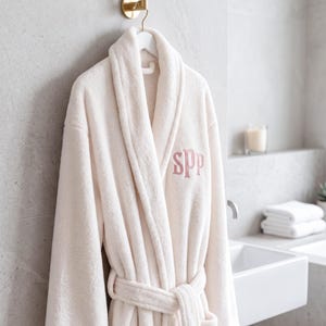 A plush marshmallow-colored fleece robe hangs elegantly on a gold hook against a gray stone wall. The robe features a personalized "SPP" monogram thêu (embroidered) on the chest in a soft flesh pink thread, showcasing a premium and serene spa-like atmosphere.