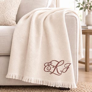 May include: A cream-coloured waffle weave throw blanket featuring a decorative fringe along the bottom edge. The blanket is embroidered with a large, dark red monogram featuring the stylised cursive letters E, R, and J in a flowing script design.