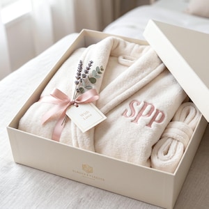 A marshmallow-toned plush robe is neatly folded inside a luxury cream gift box, decorated with a pink ribbon and a "With Love" tag. The focal point is the thick "SPP" block monogram thêu (embroidered) in flesh pink thread, emphasizing its appeal as a high-end personalized gift.
