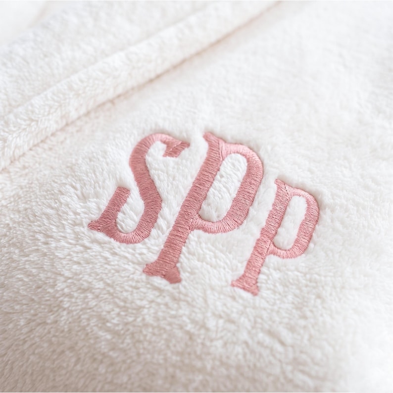 An ultra-realistic close-up shot highlighting the soft, fluffy texture of the marshmallow fleece fabric. The intricate stitching of the "SPP" monogram is clearly visible, showing the subtle contrast and high quality of the flesh pink embroidery thread.